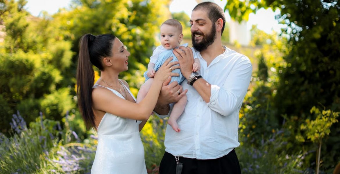 Mother and father joyfully hold their baby in a vibrant outdoor garden setting.