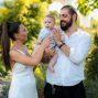 Mother and father joyfully hold their baby in a vibrant outdoor garden setting.