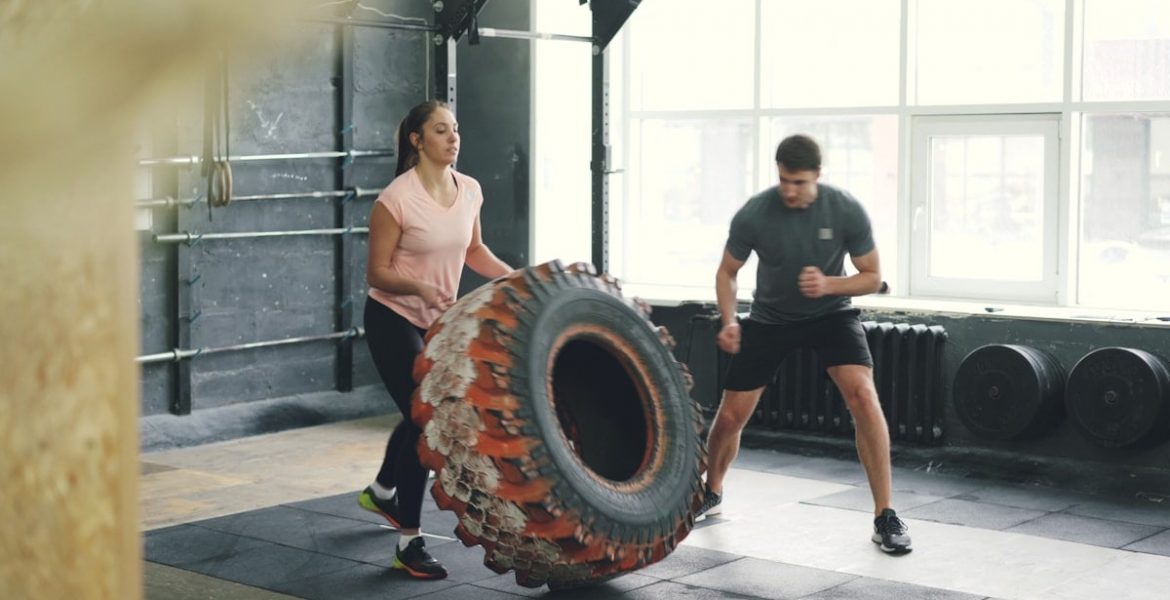 Girl in sports outfit is lifting and flipping heavy rubber tyre working out with coach in gym enjoying sports training together. People and activity concept.
