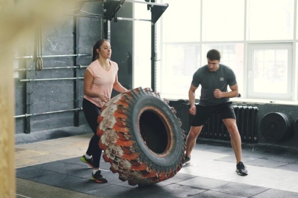 Girl in sports outfit is lifting and flipping heavy rubber tyre working out with coach in gym enjoying sports training together. People and activity concept.