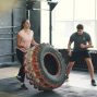 Girl in sports outfit is lifting and flipping heavy rubber tyre working out with coach in gym enjoying sports training together. People and activity concept.